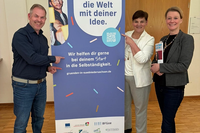 Drei Personen stehen neben einem Aufsteller. Auf dem Aufsteller steht 