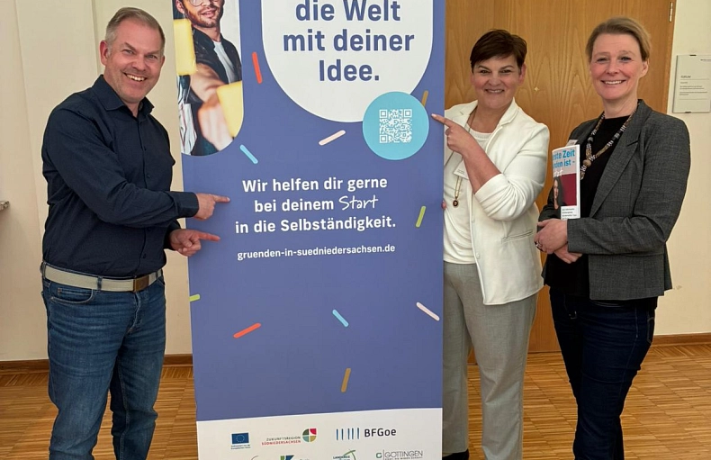2026-05-13 PM Sprechtag NBank und Gründungsnetzwerk Drei Personen stehen neben einem Aufsteller. Auf dem Aufsteller steht "Gründungsnetzwerk Südniedersachsen. Verändere die Welt mit deiner Idee. Wir helfen dir gerne bei deinem Start in die Selbstständigkeit. gruenden-in-suedniedersachsen.de". Der Aufsteller hat außerdem grafische Elemente wie verschiedene Logos.