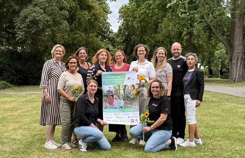 2026-07-16 PM Aktionswoche der pflegenden Angehörigen Eine Gruppe Menschen steht in einem Park auf einer Rasenfläche. Zwei von ihnen halten ein Plakat mit der Aufschrift "Aktionswoche der pflegenden Angehörigen 04.10. - 10.10.2026". einige Personen halten Sonnenblumen. Im Hintergrund sind Bäume.