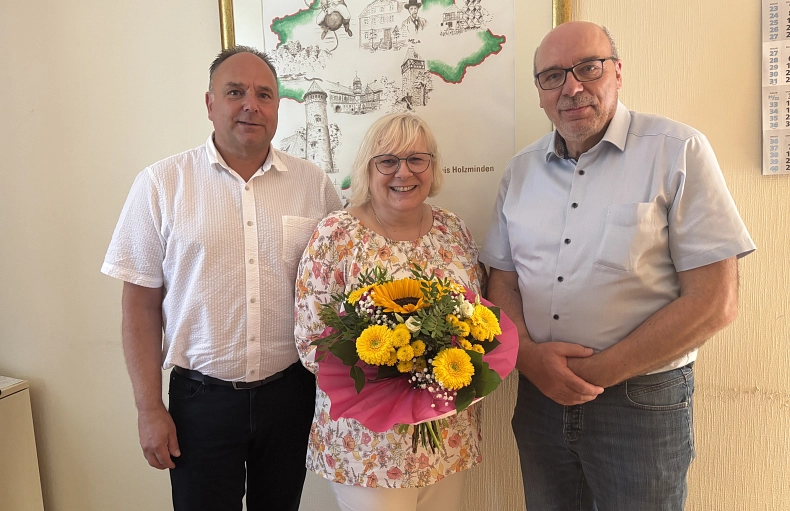 PM Abschied nach 44 Jahren Eine Frau steht mit einem Blumenstrauß zwischen zwei älteren Männern. Alle lächeln.