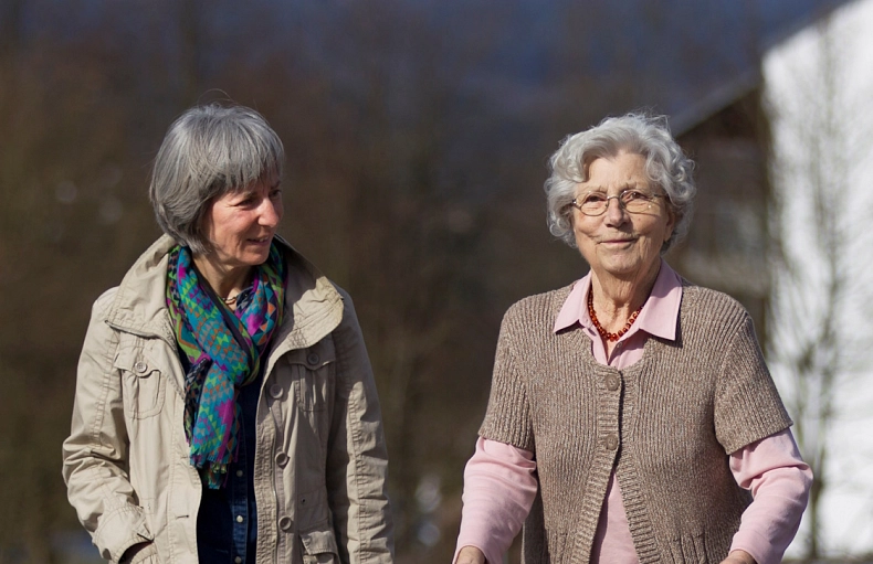 Seniorenbegleitung Eine Frau läuft neben einer älteren Frau, die einen Rollator hat. Beide sind in guter Stimmung und Lächeln. Im Hintergrund sieht man Bäume.